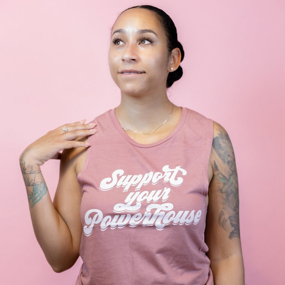Woman standing against a pink  background wearing a mauve tank top that says "support your powerhouse"