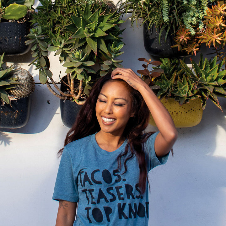 Woman wearing a blue shirt that says "Tacos. Teasers. Top Knots." in black writing