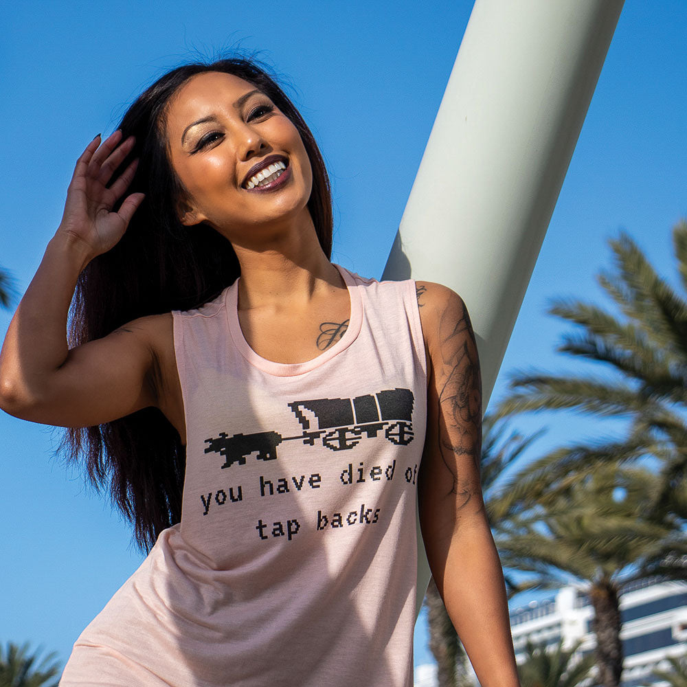 Woman wearing a women's cut peach muscle tank top that says you have died of tap backs under an old school Oregon Trail Covered Wagon in black text