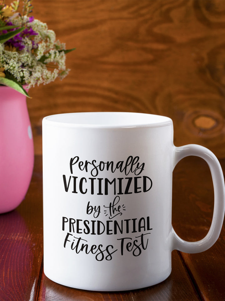 11 oz white ceramic mug that says "Personally victimized by the presidential fitness test"