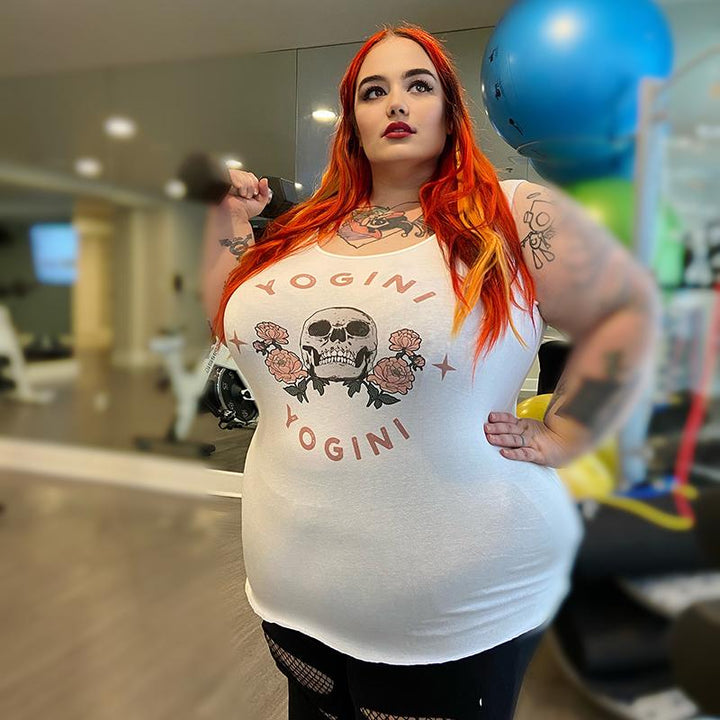Woman wearing a white muscle tank top with the words "Yogini" in a semi circle on the top and bottom of the design. There is a skull in the middle of the tank top.