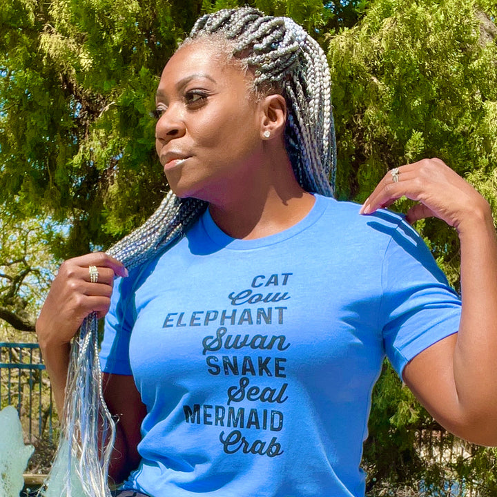 Woman wearing a unisex heather columbia blue t-shirt from The Movement Shop that says Cat, Cow, Elephant, Swan, Snake, Seal, Mermaid, Crab