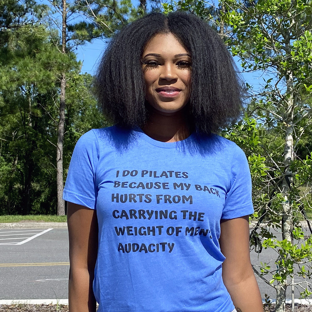 Woman wearing a blue t-shirt that says "I do Pilates because my back hurts from carrying the weight of men's audacity". T-Shirt is from The Movement Shop