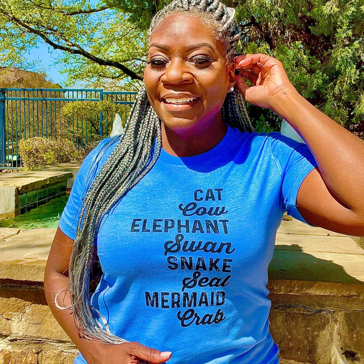 Woman wearing a unisex heather columbia blue t-shirt from The Movement Shop that says Cat, Cow, Elephant, Swan, Snake, Seal, Mermaid, Crab