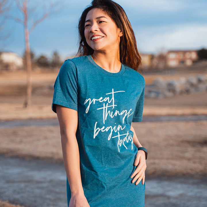 Woman wearing a unisex crewneck deep heather teal t-shirt that says "great things begin today"  in white script text