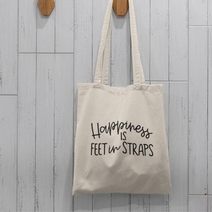 Canvas tote bag hanging on a wooden hook against white wood background. The front of the pilates tote bag reads "Happiness Is Feet In Straps" in black cursive
