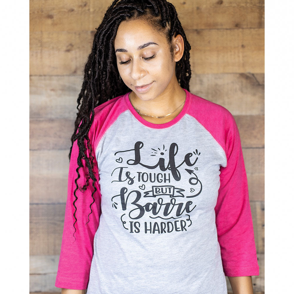 Woman leaning against a wood grain backdrop wearing a 3/4 sleeve unisex baseball shirt with pink arms and neck and grey body. Text on the front of the shirt says "Life is tough but barre is harder"