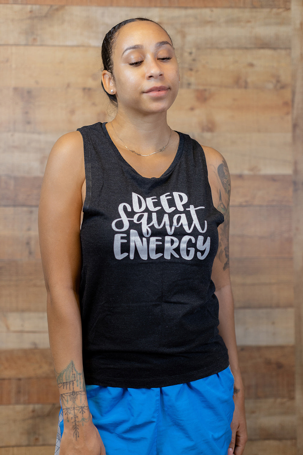 Black muscle tank top with 1" armhole that says "Deep Squat Energy" on the chest in white font. Woman leaning against a wooden background.
