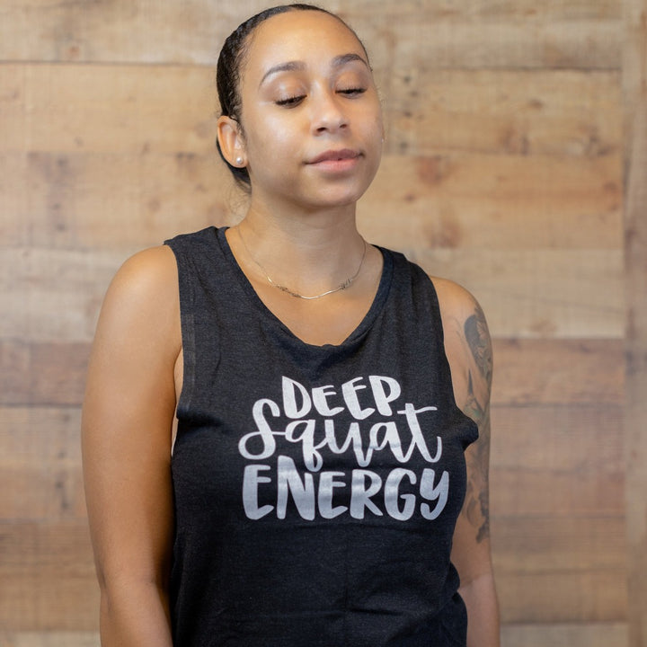 Black muscle tank top with 1" armhole that says "Deep Squat Energy" on the chest in white font. Woman leaning against a wooden background.