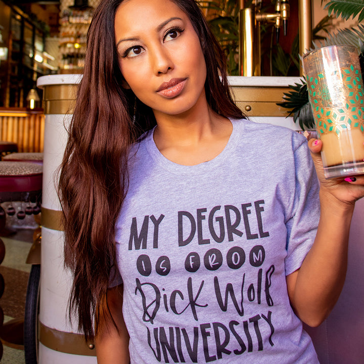 Woman wearing a heather columbia unisex crew neck blue t-shirt that says "my degree is from dick wolf university" in black text