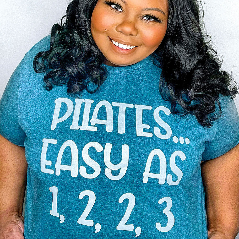 Woman wearing a heather deep teal unisex crewneck t-shirt that says "Pilates easy as 1,2,2,3" in white text