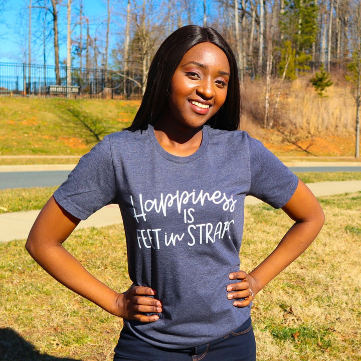 A woman wearing a heather navy unisex crewneck t-shirt that says "happiness is feet in straps" in white script text 