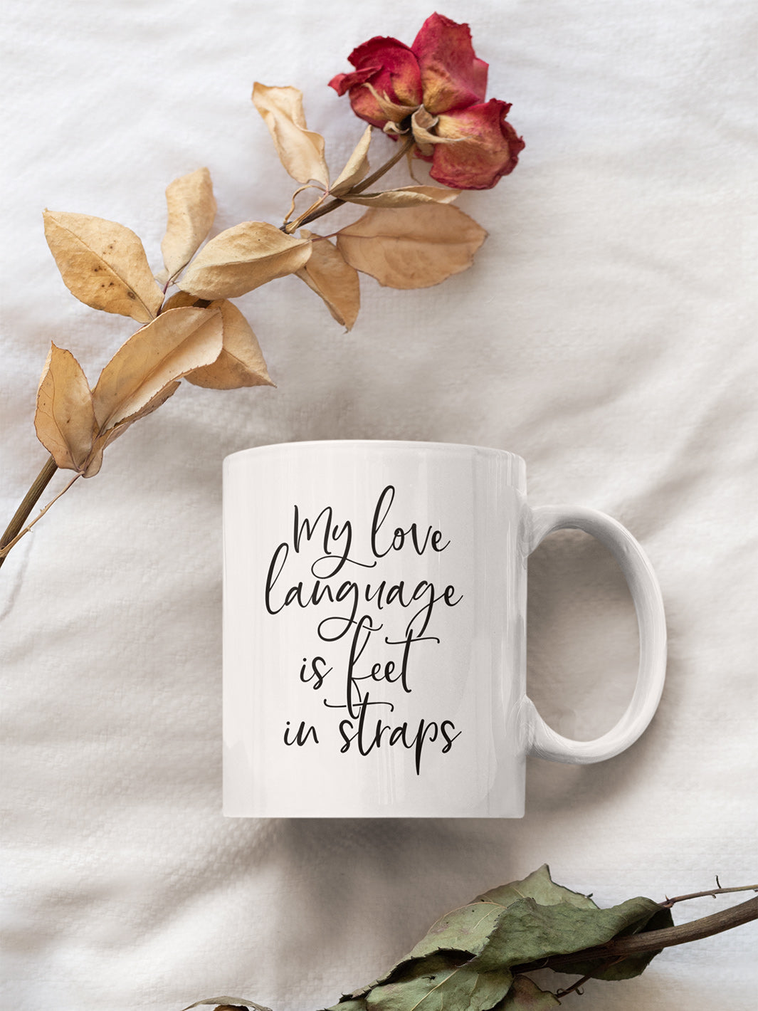 White 11oz coffee mug against a white background with dried roses that said "My Love Language Is Feet In Straps" black script  