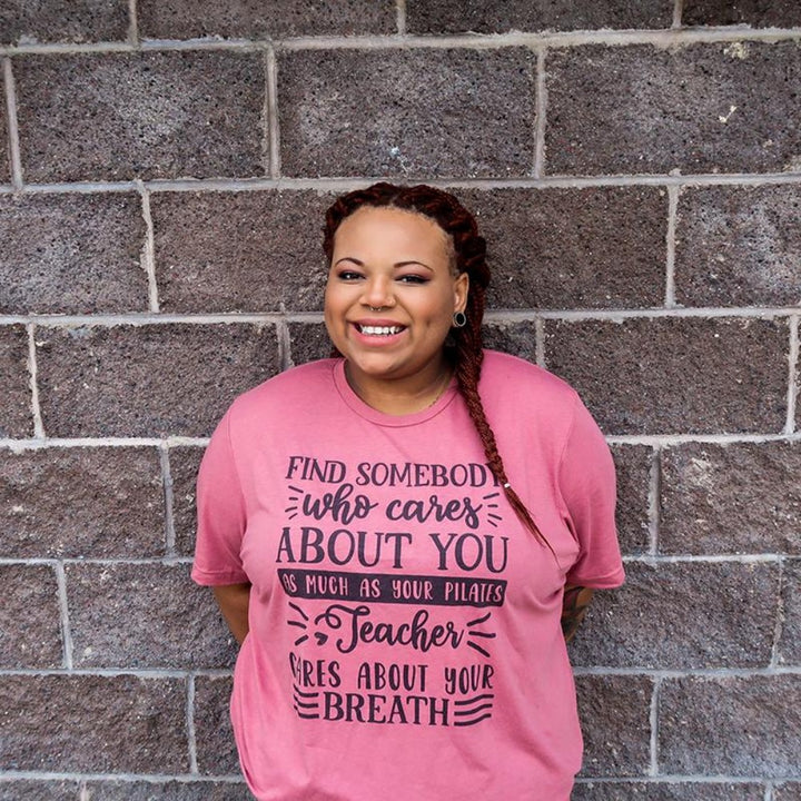 Woman wearing a unisex crewneck heather mauve t-shirt that says "Find Somebody Who Cares About You As Much As Your Pilates Teacher Cares About Your Breath" 