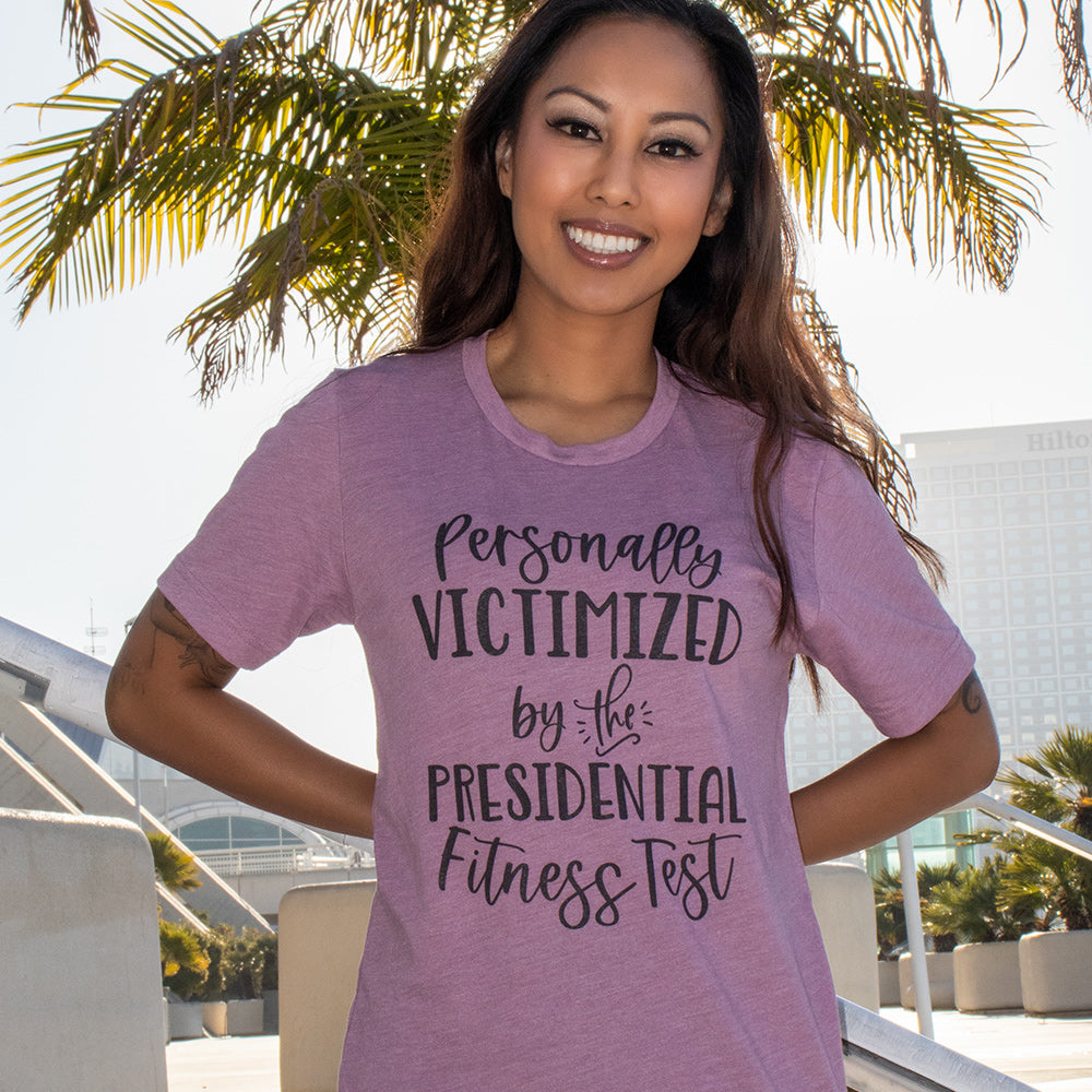 A woman wearing a heather orchid unisex crewneck t-shirt that says "Personally Victimized by the president fitness test"