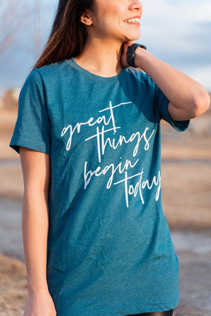 Woman wearing a unisex crewneck deep heather teal t-shirt that says "great things begin today"  in white script text
