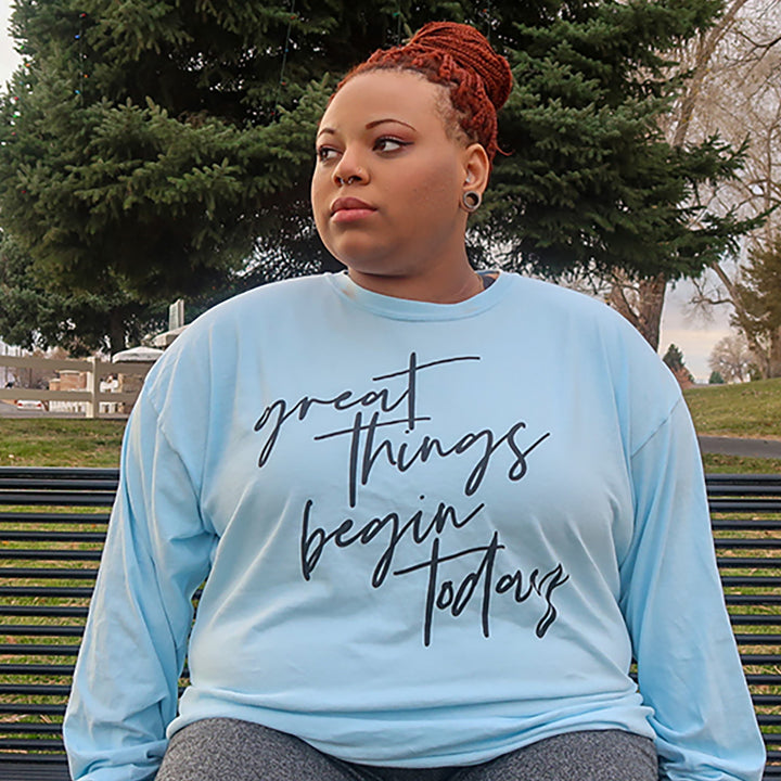 Woman wearing a light blue long sleeve 100% cotton shirt that says "great things begin today" in black script
