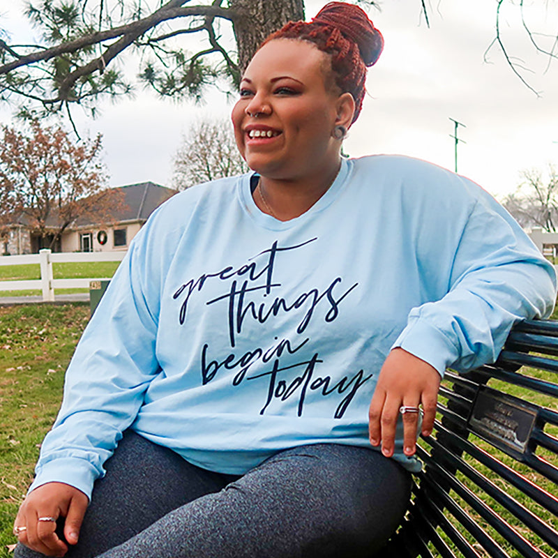 Woman wearing a light blue long sleeve 100% cotton shirt that says "great things begin today" in black script