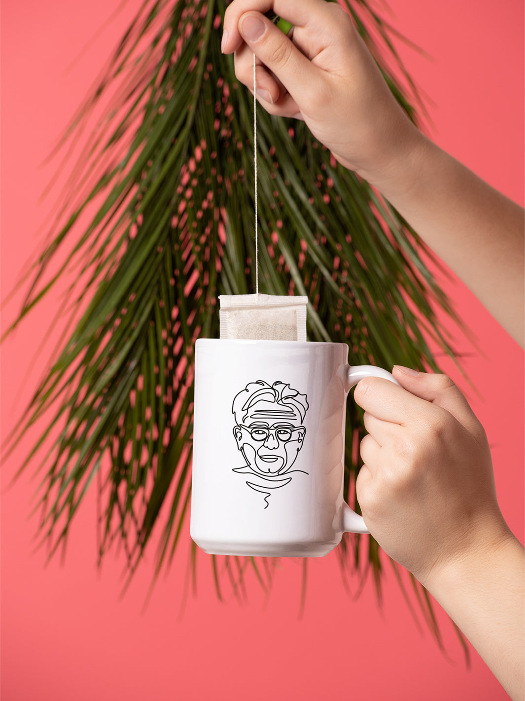 Single Line Drawing Of Joseph Pilates on a 15oz white coffee mug. There is a woman's hand dunking a tea bag against a coral background with a palm from.