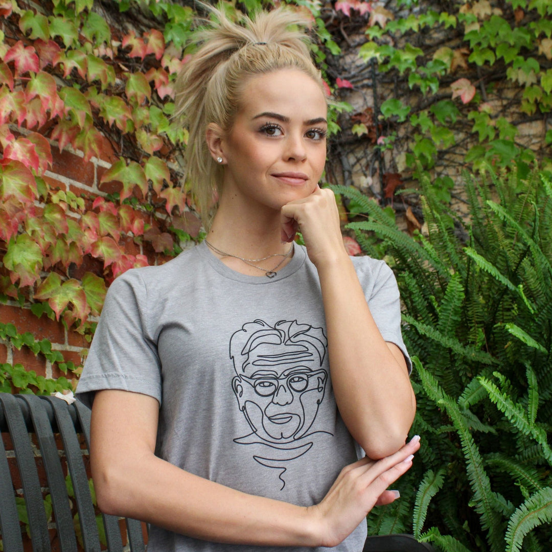 A woman wearing a grey unisex crewneck t-shirt with a line drawing of Joseph Pilates on the front. Joseph Pilates is drawn in black. 