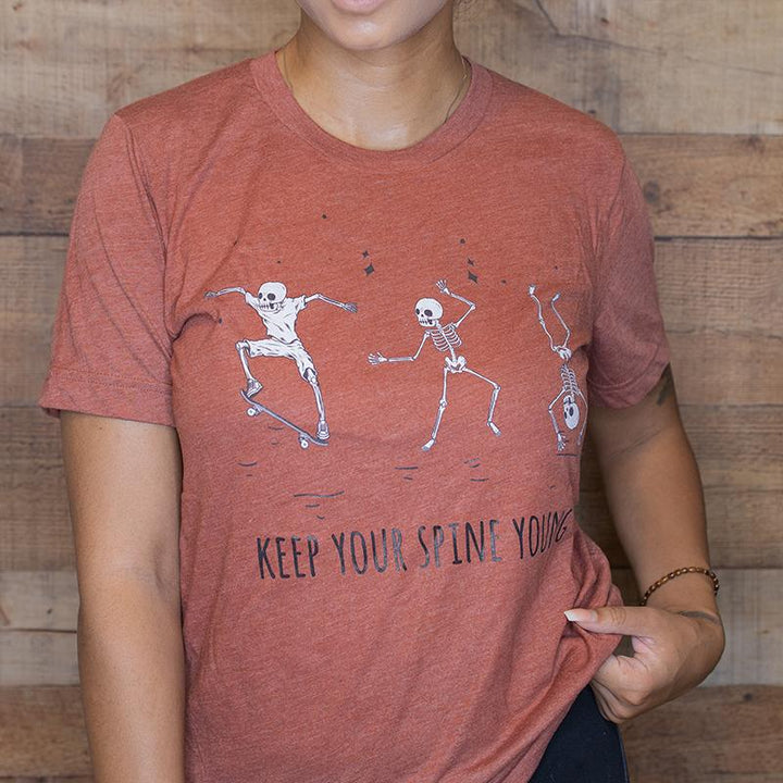 Woman wearing a heather orange unisex crewneck t-shirt that has three skelatons dancing. Text under the skelatons says Keep Your Spine Young in black text.