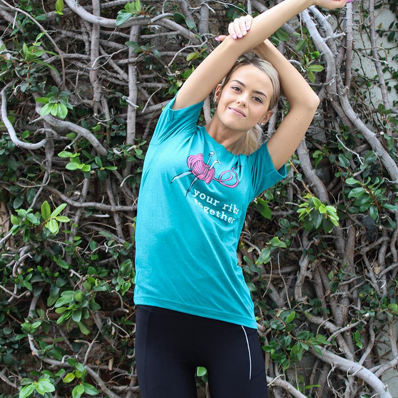 Woman wearing a deep heather teal unisex Crewneck T-Shirt that has a deep purple knitting needles and ball of yarn with the words "your ribs together:" in white text. 