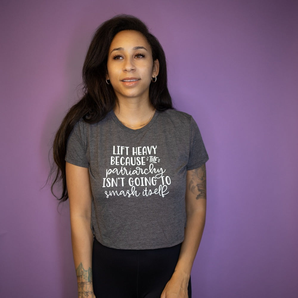 Woman wearing a deep heather crop top tshirt that says "Lift Heavy Because The Patriarchy Isn't Going To Smash Itself". Woman is leaning against a purple background.