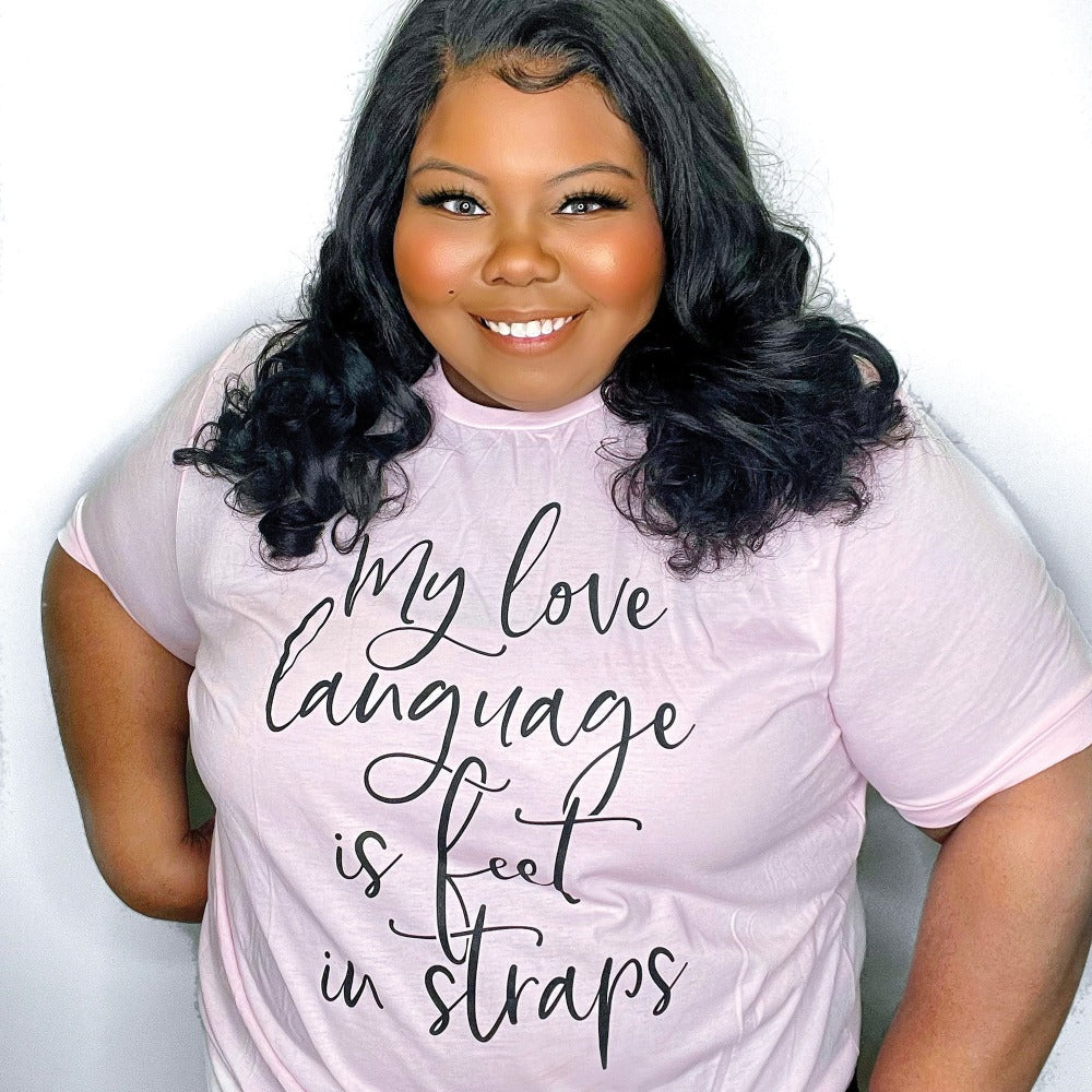 Woman wearing a heather pink unisex crewneck t-shirt that says "My Love Language Is Feet In Straps" in black script