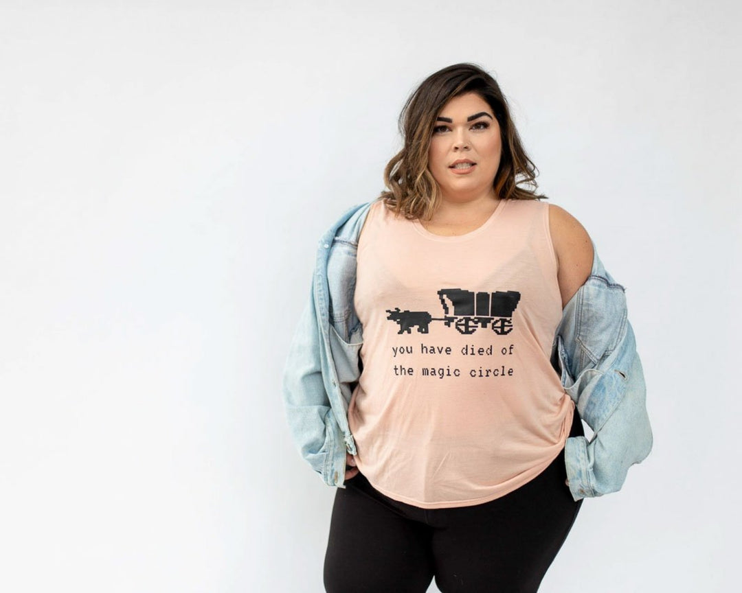 Woman wearing a peach muscle tank that says "You have Died of the Magic Circle in black text with the Oregon trail covered wagon