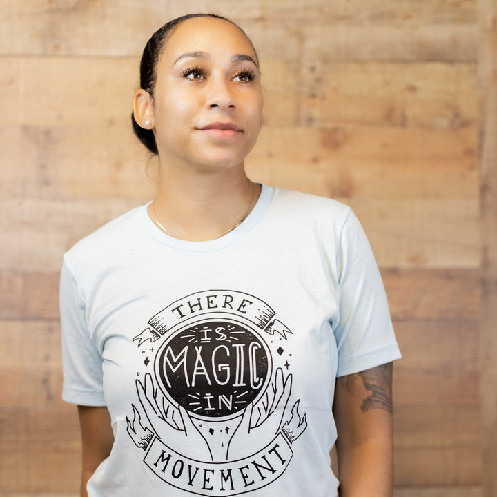 woman wearing heather ice blue crewneck t-shirt that says "There Is Magic In Movement" in black text. The words "is Magic In" is designed to look like a crystal ball. 