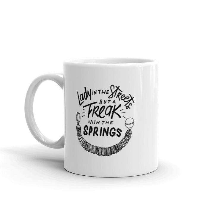11oz Coffee Mug that says Lady In The Streets But A  Freak With The Springs. There is Pilates reformer spring under the text. Design is in black.