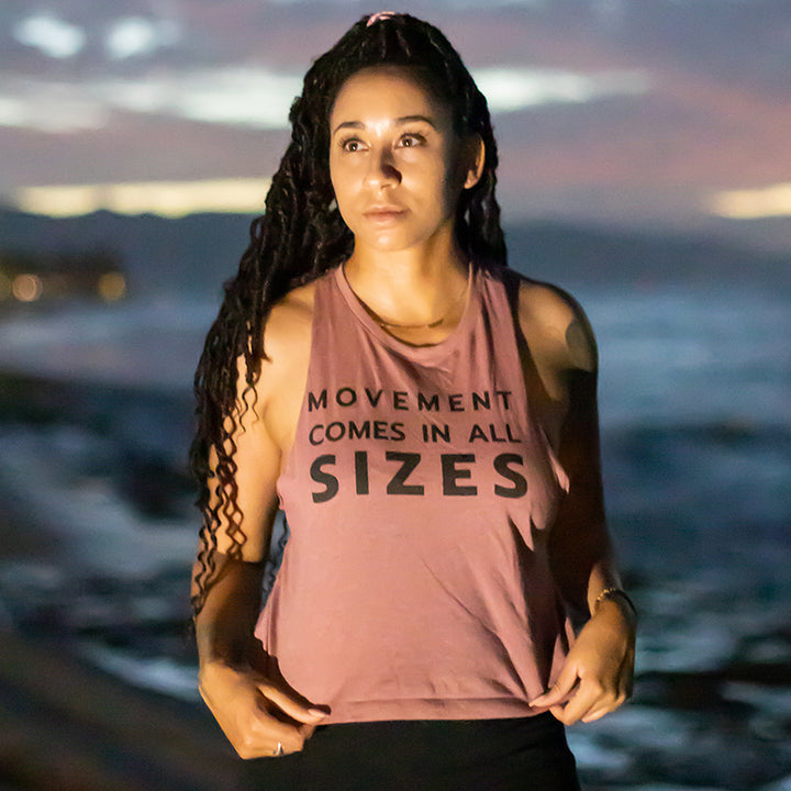 Woman wearing a mauve crop muscle tank top that says "Movement Comes In All Sizes". Against a sunset beach background.