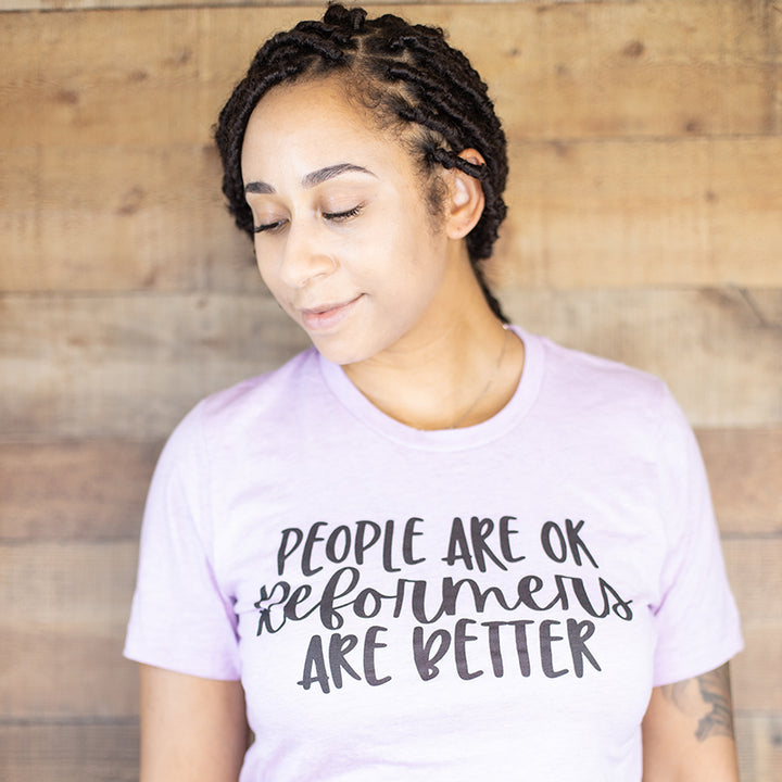 Woman leaning against a wooden background while wearing a shirt that says "People Are Ok, Reformers Are Better" in black text