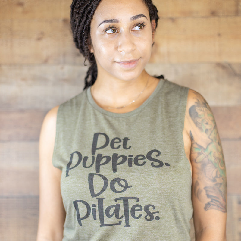 Woman wearing a heather olive woman's cut muscle tank top that says Pet Puppies. Do Pilates. in black text. 
