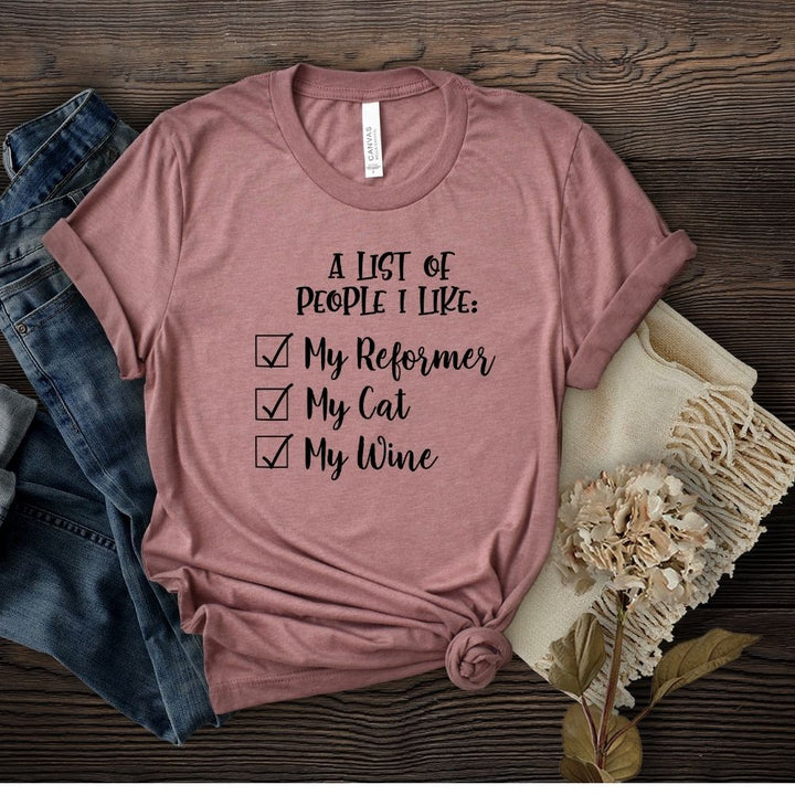 A mauve unisex crew neck t-shirt that says "A list of people I like: My reformer, A Cat, my wine". Mauve shirt is against a wooden backdrop with flower and jeans props.