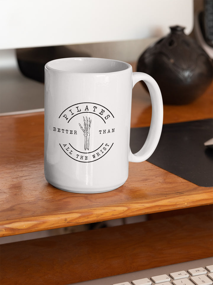 White 15oz mug with handle on the right. The mug says "Pilates Better Than All The Wrist" with a skeleton hand in black text. The mug is on a wood desk. 