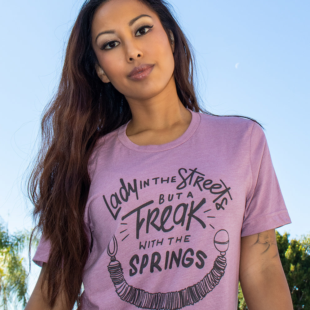 Woman wearing a heather orchid crew neck t-shirt that says "Lady in the streets but a freak with the springs" and a Pilates Reformer Spring design
