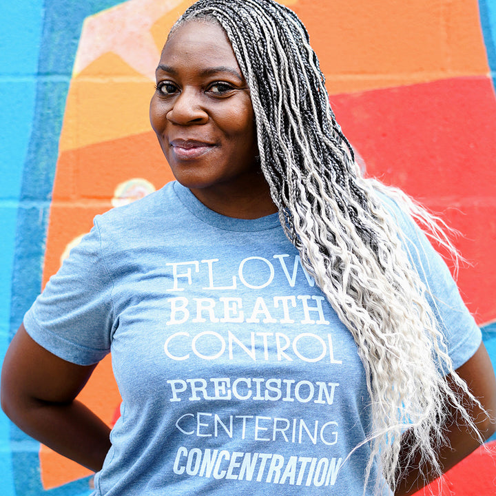 Woman wearing a blue unisex crew neck t-shirt that lists the 6 Pilates Principles "Flow, Breath, Control, Precision, Centering, Concentration"