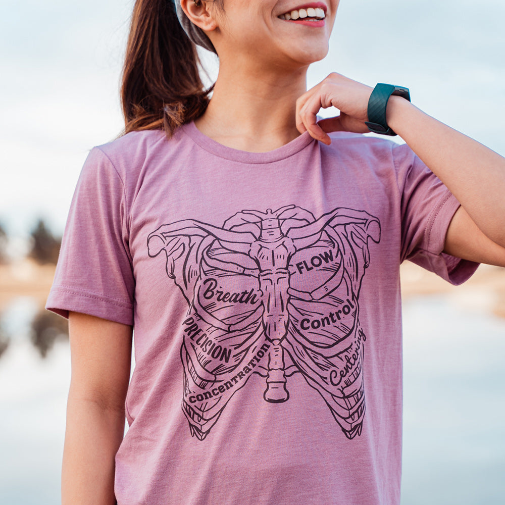 Woman wearing a heather orchid t-shirt with a drawing of a rib cage with the ribs designed as the 6 pilates principles 