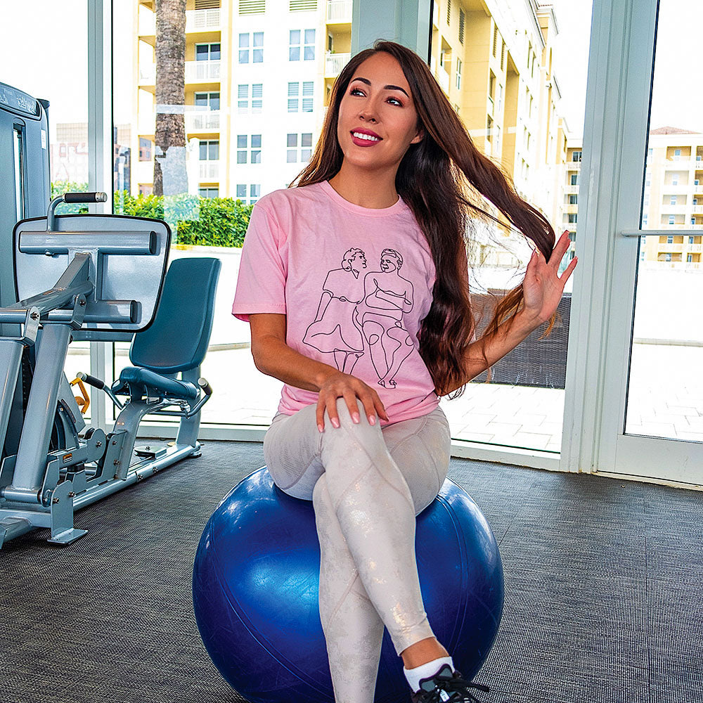 A woman sitting on a blue exercise ball wearing a unisex crew neck t-shirt that is a single line drawing of Clara Pilates and Joseph Pilates