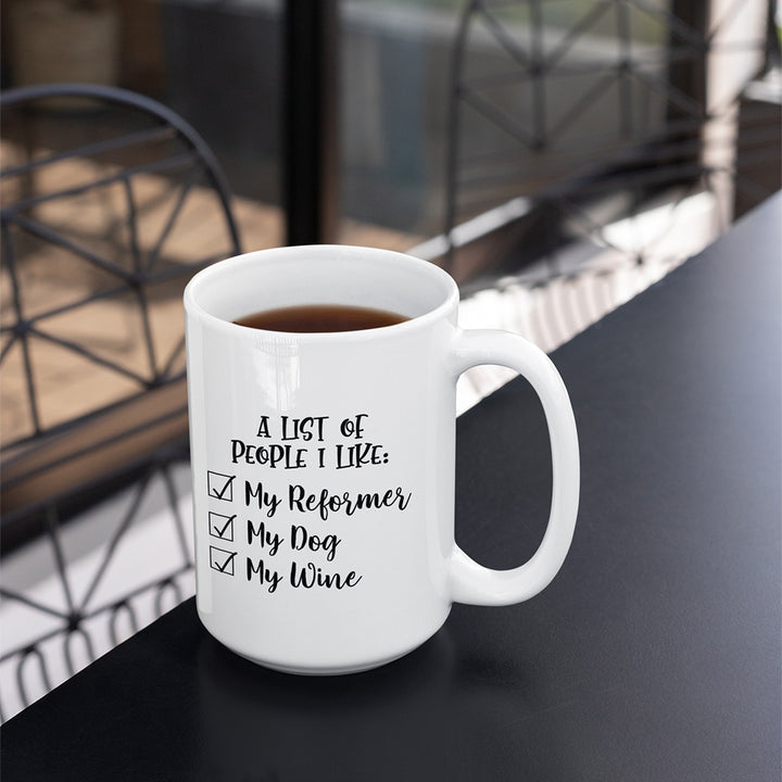 15 oz white coffee mug that says "A list of people I like: My reformer, My dog, My Wine". Coffee mug is sitting on a black table with chairs in the background. 