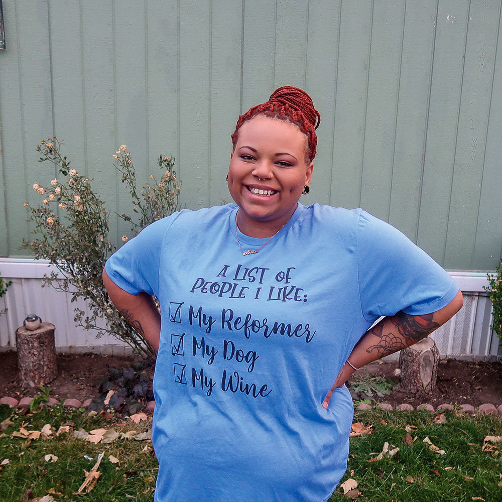 Woman wearing a heather columbia blue unisex crewneck t-shirt that says "A list of people I like: my reformer, my dog, my wine" in black script