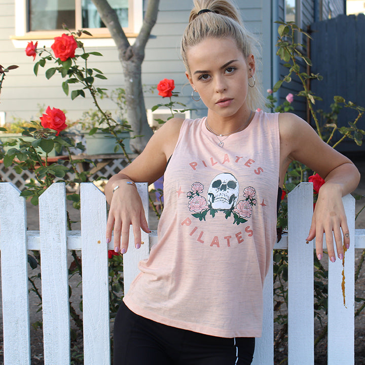 Woman wearing a peach muscle tank with a skull on it and the words Pilates over and under the skull. Woman is leaning against a white fence.