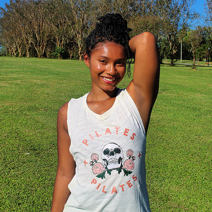 Woman wearing a white muscle tank with a skull on it and the words Pilates over and under the skull.