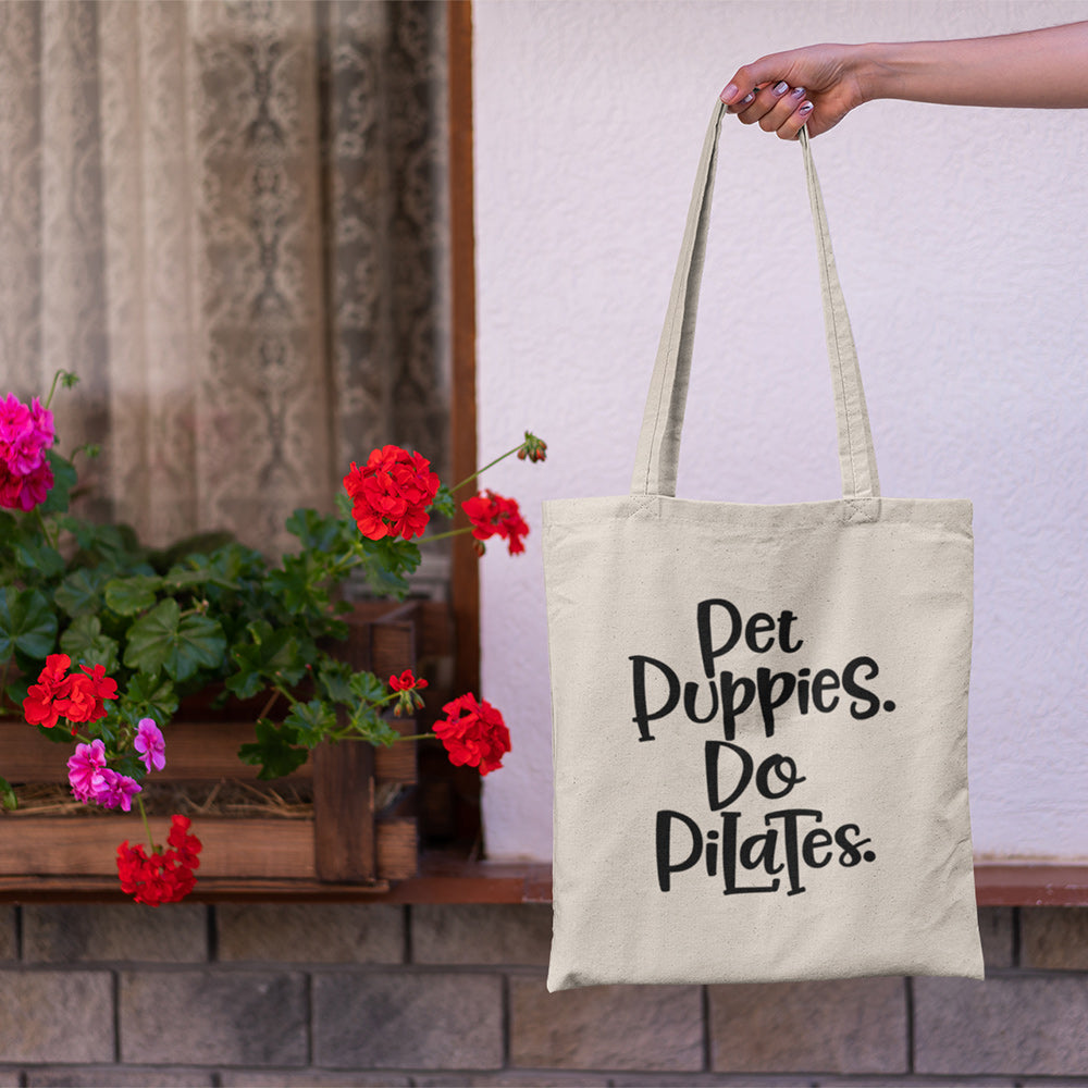 Outstretch hand holding a canvas tote bag by it's handles. On the tote bag reads "Pet Puppies. Do Pilates."