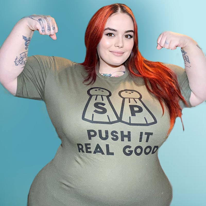 Woman wearing a heather olive unisex crew neck t-shirt that says "Push It Real Good" and has two salt and pepper shakers