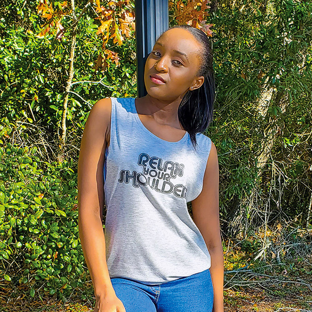 Woman leaning against a pole with a nature inspired background. Woman is wearing a grey tank top that says "Relax Your Shoulders" in black retro font.