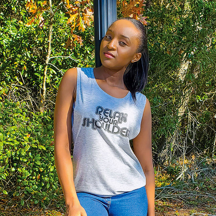 Woman leaning against a pole with a nature inspired background. Woman is wearing a grey tank top that says "Relax Your Shoulders" in black retro font.