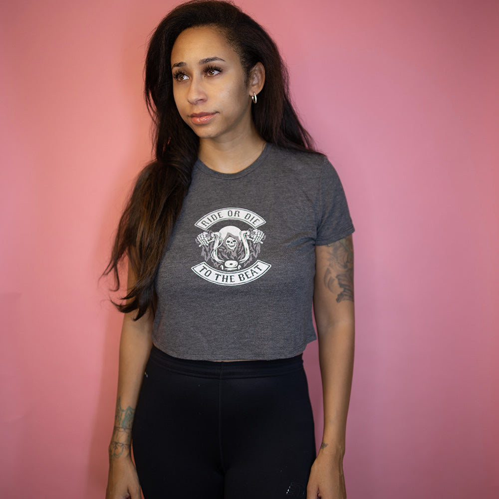 Woman wearing a crop top t-shirt that says "Ride Or Die To The Beat" in white text. Woman is leaning against a pink background