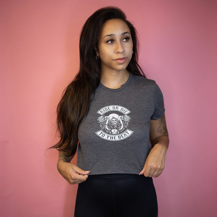 Woman wearing a crop top t-shirt that says "Ride Or Die To The Beat" in white text. Woman is leaning against a pink background